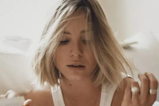 Blondynka z fryzurą bubble bob, zatopiona w lekturze, otoczona jasnymi, miękkimi tkaninami.