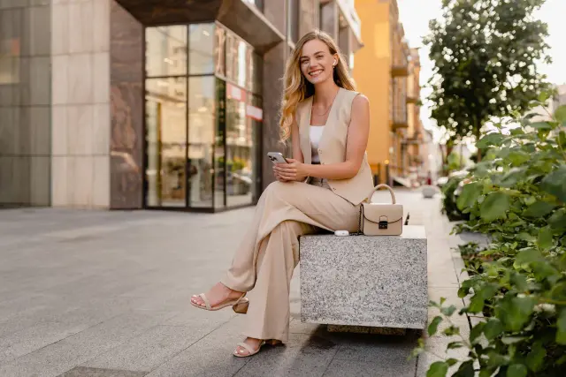 Uśmiechnięta kobieta w beżowym garniturze, idealnym na styl smart casual damski, siedzi na ławce z telefonem w dłoni.